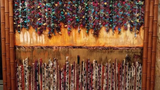 Enclosed in a thick bamboo-style frame, the piece is divided horizontally. The top section features a dense, vibrant canopy of clustered textile elements hanging over a textured, golden-orange background. The bottom section consists of dense, vertical rows of multicolored bound threads standing upright.The bottom section consists of dense, vertical rows of multicolored bound threads standing upright.