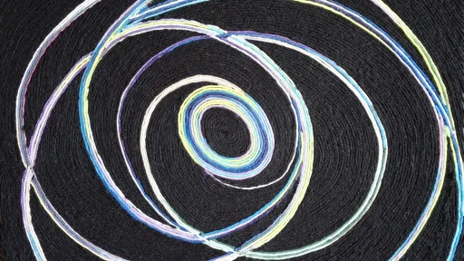 The piece features multicolored woolen threads—in shades of blue, teal, purple, yellow, and white—arranged in intersecting, continuous circular patterns against a solid, textured black background. The threads form concentric and overlapping rings that radiate from a tight central spiral, resembling planetary orbits or swirling cosmic rings.