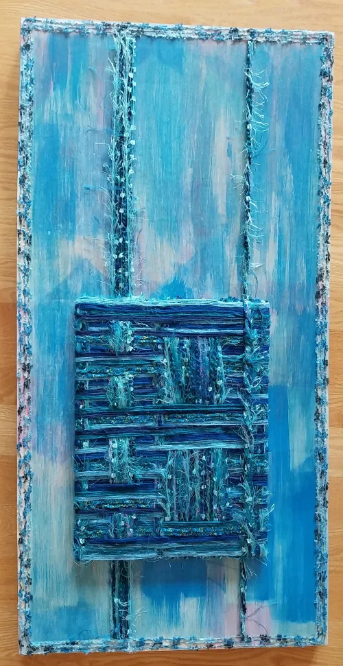 The piece features a painted abstract background in fluid shades of light and dark blue acrylic, resembling water. Overlaid in the center is a heavily textured, woven square made of interlaced blue, teal, and green threads forming a grid pattern. Two vertical bands of similar frayed threads run from the top to the bottom of the canvas, passing beneath the central square. The artwork is framed by a thin, fuzzy border of textured blue thread.